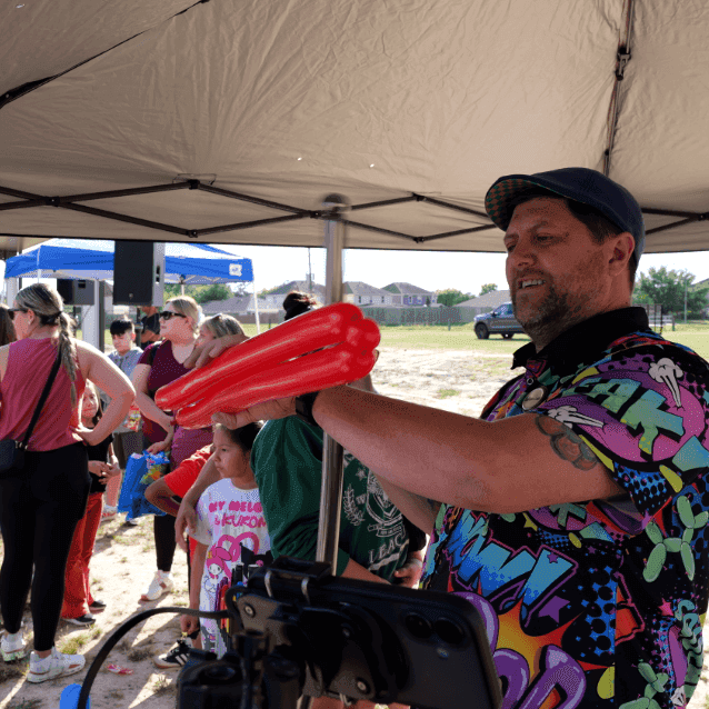 A balloon artist in a colorful shirt creates a red balloon animal for children waiting in line under a tan canopy at an outdoor event.
