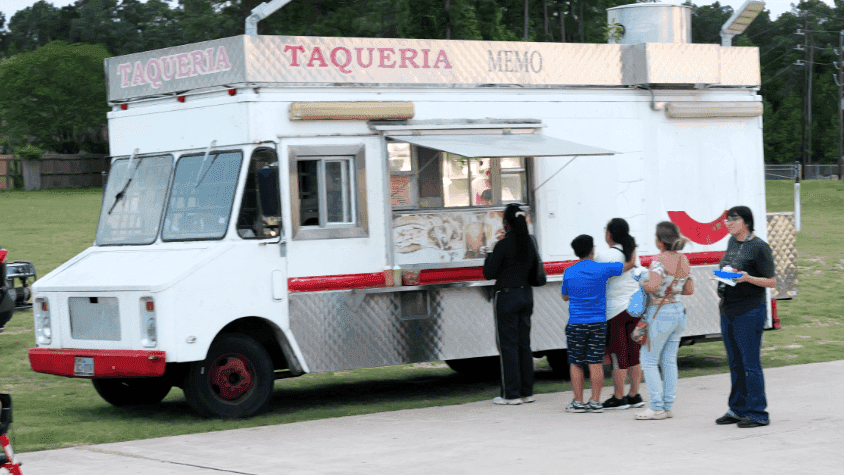 Residents stand in line to order food from the window of a large white Taqueria Memo food truck parked on a grassy field at dusk.
